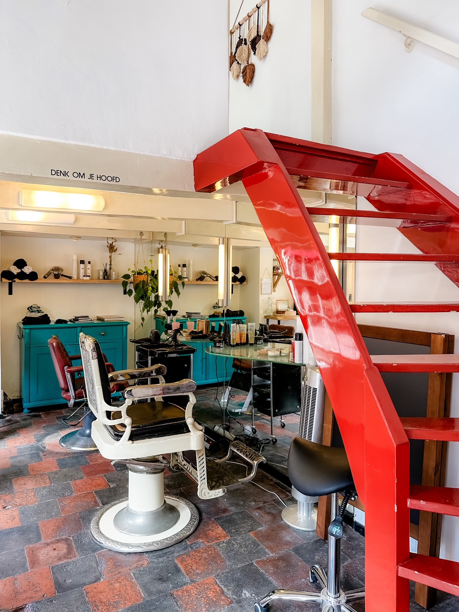 De salon van binnen: een rode wenteltrap, een witte kappersstoel, kasten in petrol en een vloer van rode en grijze tegels