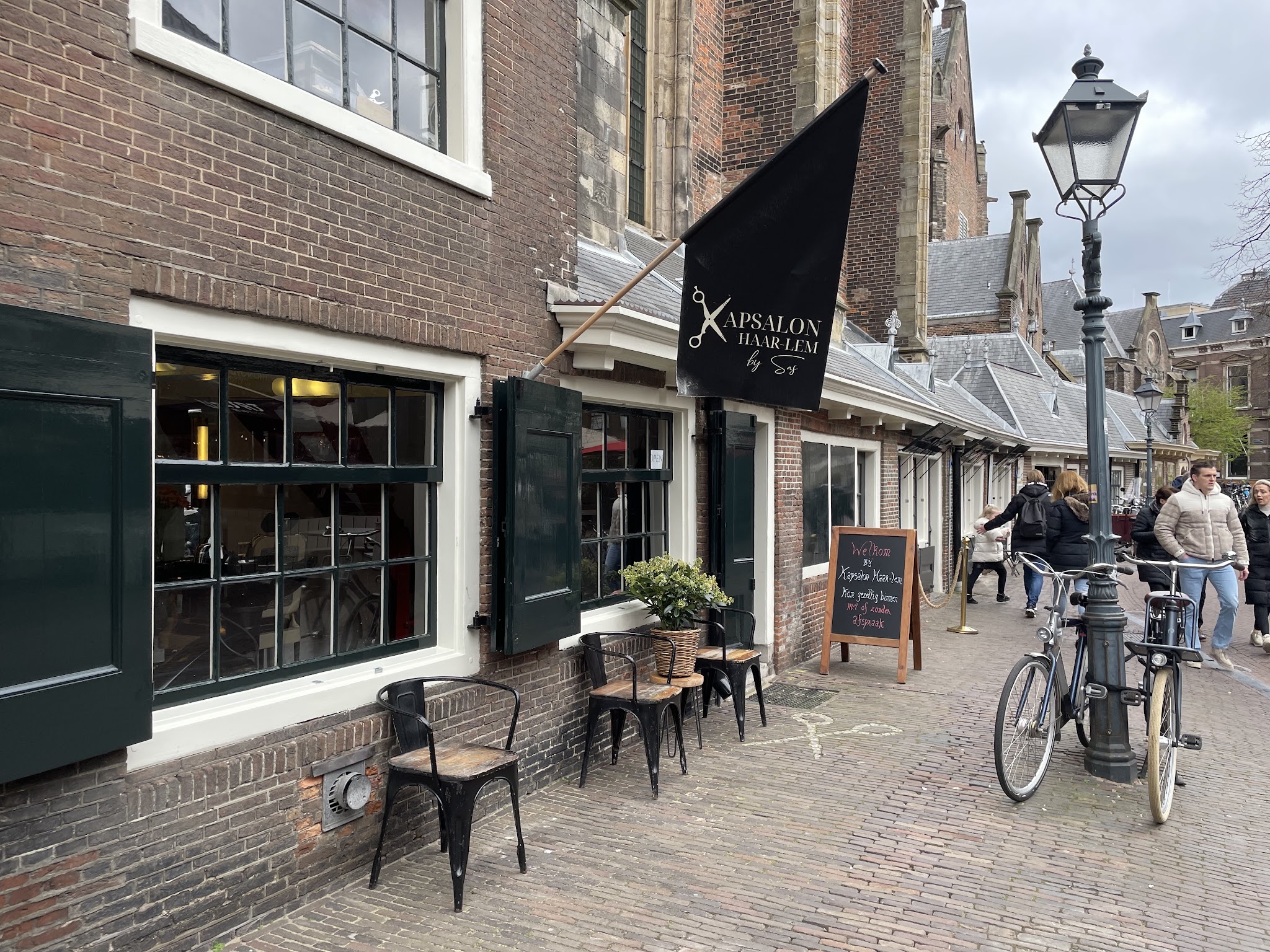 De gevel van Kapsalon Haar-lem aan de Oude Groenmarkt, met de kleine raampjes, de zwarte vlag, een lantaarnpaal en stoeltjes op de stoep