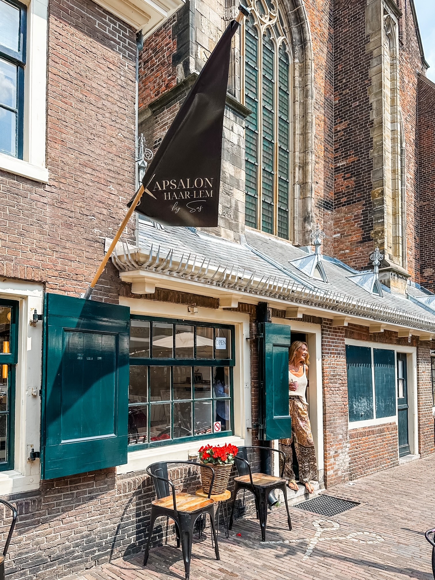 De voorkant van Kapsalon Haar-lem tegen de muur van de kerk, met de zwarte vlag boven de deur, groene luiken en stoeltjes met rode bloemen op de stoep