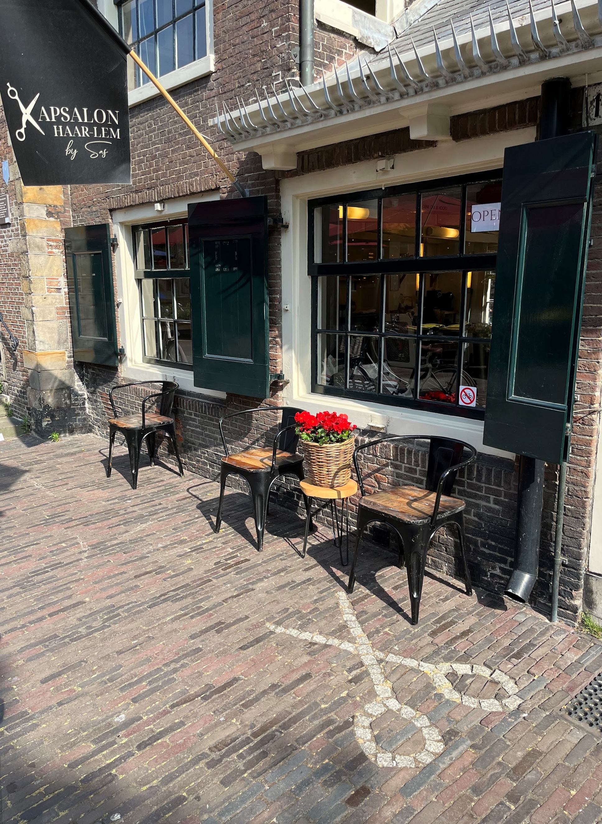 De gevel van Kapsalon Haar-lem aan de Oude Groenmarkt, met de zwarte vlag, groene luiken, stoeltjes op de stoep en een schaar van witte steentjes in de bestrating